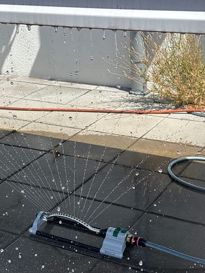 The media file depicts a close-up view of a sprinkler system in operation on a paved outdoor surface. The sprinkler itself is a black and gray device with a water hose attached to its top. It is positioned on dark gray paving stones, and a fan of water is being sprayed outwards, creating a pattern of fine droplets. Some of these droplets are visible in the air and on the ground. Adjacent to the dark pavers is a section of lighter, beige-colored pavers, also showing water droplets.
To the left of the paved area, there is a vertical white structure, possibly a wall or a frame. On top of this white structure, and extending over the scene, is a dry, brown, leafy plant. Water is also visible dripping down the side of this white structure, forming small rivulets and individual drops. A thin orange hose or wire runs vertically between the white structure and the pavers.
The lighting suggests it is daytime, with shadows cast on the paving stones, indicating the sun is present. The weather appears to be clear, and the scene is likely outdoors, possibly in a garden or patio area. The focus is on the water and the mechanics of the sprinkler, suggesting an activity of watering plants or cooling down an area. There is no visible text in the image. The overall impression is one of a functional, possibly domestic, outdoor setting.
