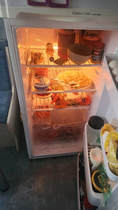 This image shows an open, brightly lit refrigerator, likely in a kitchen or dining area. The refrigerator is filled with various food items and beverages. On the top shelf, there are eggs and a package that appears to be food items. The middle shelves contain several jars, including one labeled "spilva Sinepes stiprās" (spilva Mustard strong) and another that seems to be jam. There's also a bottle of Corona beer. A bowl of French fries sits on a plate with a spoon. Other items include tubs of "Mascarpone" and what appear to be berries. A package of hot dogs is visible on the bottom shelf, alongside other food items in plastic packaging. The interior of the refrigerator is somewhat cluttered, with some items in clear plastic containers and others loose. The lighting creates a warm, inviting glow. The overall impression is one of everyday life and the contents of a home refrigerator.