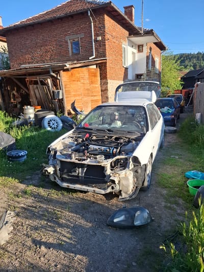 The image depicts a salvage yard in Rakitovo, Bulgaria. A partially disassembled white car, with its hood removed and engine exposed, is the central object. Next to the car on the ground is a detached headlight. The car is parked on a dirt and grassy surface, surrounded by other car parts, tires, and miscellaneous items. In the background, there are several other vehicles, including a dark-colored car, and a building with a brick facade. A wooden shed is positioned adjacent to the brick building. The sky is clear and blue.