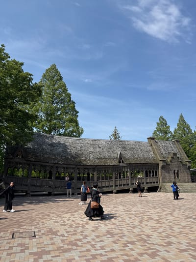 The image shows a covered walkway with dark wood lattice sides and a stone tile roof. People dressed in dark robes walk along the walkway. To the left of the walkway are green trees and a blue sky with some clouds. The ground is covered in red, brown, and beige tiles. The location is likely the Harry Potter section of Warner Bros. Studio Tour Tokyo in Nerima, Japan.