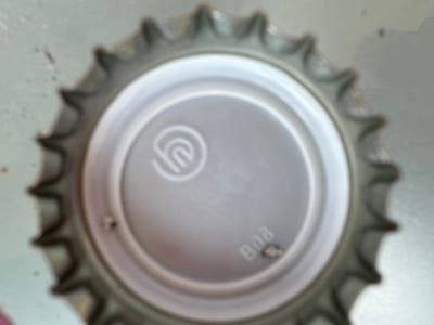 A close-up, top-down, slightly blurry photograph shows the interior of a silver bottle cap. The cap has a ridged outer edge and a smooth, concave center. Embossed in the center of the cap is a circular logo with a stylized "n" inside. To the right of the logo, the number "808" is faintly visible in a raised, embossed font. The background is a light gray, textured surface, possibly a tabletop or counter. There are a few small dark specks on the surface. The image is of a bottle cap, likely found in Villa Pasquali, Italy.