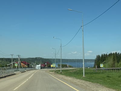 The image depicts a road in Mysy, Russia, leading towards a body of water, possibly a lake or wide river. The road is asphalt with a yellow painted line marking the edge. Streetlights line the right side of the road. Utility poles and electrical wires are visible along the left side of the road. Several buildings, some with red roofs, are visible in the distance on the left. "Pedestrian Crossing" signs are present in multiple locations. There is a forest of coniferous trees on the right. The sky is clear and blue. The weather appears to be sunny.