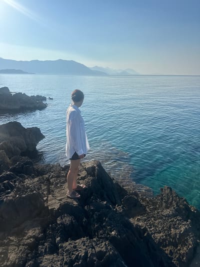 The image depicts a person standing on a rocky coastline overlooking a calm, turquoise body of water. The person, facing away from the camera and towards the water, is wearing a white oversized shirt and dark shorts. The sun is high in the sky, casting a bright light and creating a lens flare in the upper left corner. In the distance, hazy mountains rise from the horizon. The rocky shore is dark and rugged, with some areas showing submerged rocks beneath the clear water. The overall scene suggests a peaceful and serene coastal environment. The location is identified as being in the city of Kubasi, Montenegro.