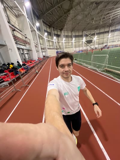 A selfie taken indoors shows a man on a red running track. He wears a white t-shirt and black shorts. A smartwatch is visible on his wrist. The background reveals an indoor stadium with seating and a netted soccer field. The photo appears to be rotated, with the ceiling and stadium structure angled to the left. The man looks toward the camera, holding his arm out for the selfie.