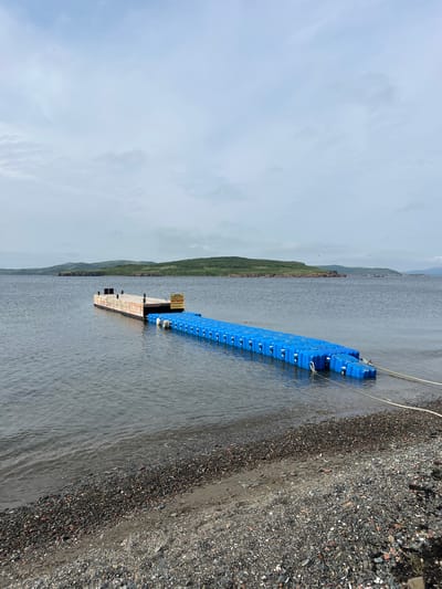 The image depicts a floating dock extending from a pebble beach into a body of water. The dock consists of a wooden section with some black bollards and a longer section made of connected blue plastic pontoons. A yellow sign with Russian text is attached to the wooden part of the dock. The water is calm, with gentle waves lapping at the shore. In the background, a hilly, tree-covered coastline stretches across the horizon under a cloudy sky. The overall environment suggests a natural, possibly remote, waterfront location. The city of Reineke, Russia, is not directly visible in the image, but the scene's characteristics are consistent with a coastal setting in that region.