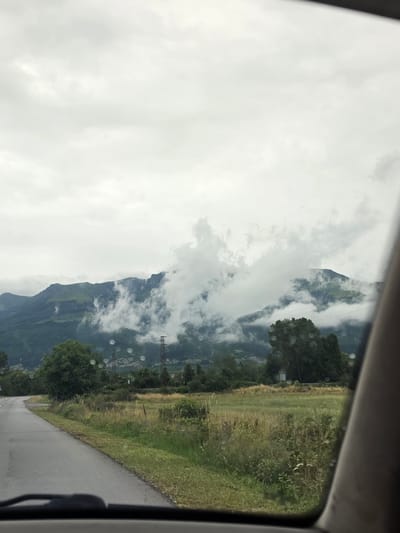 The image captures a view from inside a vehicle, looking out through the windshield. A paved road curves to the left in the foreground. To the right of the road is a grassy field with wildflowers and tall grasses. Beyond the field, several trees are visible, including a large, leafy tree to the right. In the background, a mountain range dominates the scene, with clouds obscuring parts of its peaks and slopes. Electrical towers and wires can be seen scattered across the landscape. The sky is overcast with a light gray hue. There are numerous water droplets visible on the surface of the windshield, suggesting it may have recently rained or is currently raining. No people are visible in the image. The scene appears to be a rural or semi-rural mountainous area.