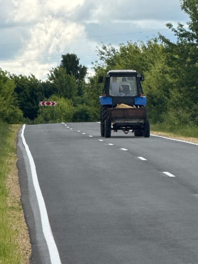 An overcast sky looms over a rural road in Steksovo, Russia. The road, paved and marked with white dashed lines, curves to the left. On the right side of the road, a blue tractor with a trailer filled with hay is traveling away from the viewer. The tractor's cabin has two visible occupants. To the left of the road, a red and white chevron sign indicates a sharp curve. Lush green trees and foliage line both sides of the road, forming a dense backdrop. The ground beside the road is covered in grass and gravel.