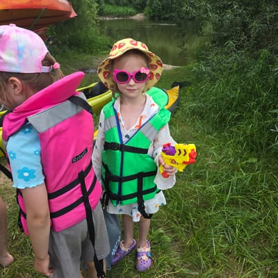 Two young girls are standing on a grassy bank next to a river. The girl on the right is wearing a straw hat with strawberries, pink cat-shaped sunglasses, a white shirt with colorful heart prints, and a green life vest. She is holding a yellow and red toy water gun. The girl on the left is wearing a tie-dye baseball cap and a pink life vest over a blue shirt with white flowers. Several kayaks are visible on the bank behind them. The river is calm and surrounded by trees. The scene appears to be a recreational area in or near Khotylevo, Russia.