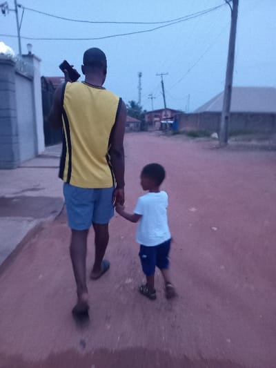 A man in a yellow and black tank top walks hand-in-hand with a young boy in a white t-shirt and blue shorts on a dirt road. The man holds a phone in his other hand. Power lines and utility poles are visible in the background. The lighting suggests it is dusk or dawn.