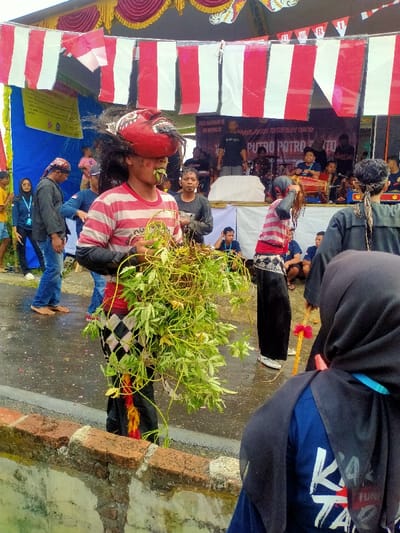 An outdoor public event is depicted in Tamansari, Indonesia. The scene is dominated by a male individual in the foreground, facing slightly right, wearing a red and white horizontal striped long-sleeve shirt, black pants, and a black and white diamond-patterned sarong-like garment over the pants. A large, red, stylized mask with dark, wild hair is positioned on his forehead, obscuring the upper part of his face. He is holding a substantial bundle of green leafy vines and branches in front of his torso, with some green foliage visibly protruding from his mouth. His feet are bare, standing on a wet, dark paved surface.
Behind this individual, to the left, are several bystanders, some observing, one male in blue jeans is also barefoot. Directly behind the main subject, another male in a dark t-shirt looks forward. To the right, slightly further back, a person with a similar red and white striped shirt and black pants is depicted with their head bowed, long hair obscuring their face, seemingly in a performance.
In the background, a temporary stage is erected, featuring a white front and a backdrop banner with "PUTRO POTRO" and other Indonesian text. Musicians are visible on the stage with traditional instruments, including drums. Overhead, red and white striped fabric banners are strung across the width of the stage area, with a small Indonesian flag visible on the left. The ground is wet, suggesting recent rain or damp conditions. In the extreme foreground on the right, the back of a person wearing a black hijab and a blue shirt with partial white lettering is visible, positioned behind a low, weathered brick wall.