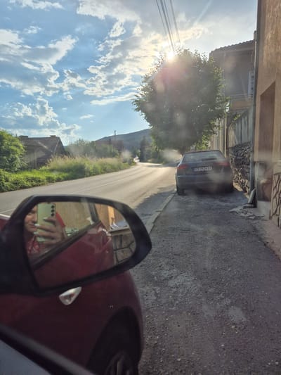 The image captures a street scene in Iskrets, Bulgaria, on a bright, sunny day. The sun is prominently visible in the upper right portion of the frame, casting a strong lens flare and illuminating the sky, which is adorned with scattered clouds. A tree with lush green foliage is positioned in the mid-ground, also illuminated by the sun.
In the foreground, a red car is partially visible, with its side mirror reflecting a woman holding a smartphone. The woman is looking at her phone, and her face is partially visible in the reflection. Her fingernails appear to be painted a light green color.
On the right side of the street, a dark-colored Audi A3 is parked, its license plate reading "CO 2124 BX." The car is close to a stone wall that runs alongside the sidewalk. The sidewalk itself is made of gravel and asphalt, with some debris scattered on it. A building with a brown facade and a tiled roof is situated behind the stone wall.
The street itself is paved and curves slightly into the distance. On the left side of the street, there are houses with red tiled roofs and green vegetation. The landscape beyond the houses appears hilly and covered with trees. Power lines are visible stretching across the sky, adding to the urban infrastructure context. The overall impression is that of a quiet, residential street in a town during daylight.