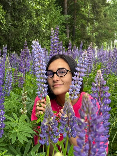 In Vakhrino, Russia, a woman with dark hair and glasses poses amidst a dense field of blooming purple lupines. She is wearing a bright pink t-shirt and is holding several lupine stalks in her hands. The background consists of lush green trees and more lupine flowers, creating a vibrant natural setting. The lighting suggests it is daytime.