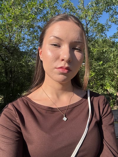 A young woman with long, brown hair and fair skin is captured in a close-up selfie outdoors. She is wearing a brown, long-sleeved shirt and a delicate gold necklace with a clover-shaped pendant. A white strap, likely from a bag, is visible across her chest. Sunlight highlights her face, creating a luminous glow. The background consists of lush green trees and a bright blue sky, suggesting a sunny day. The specific location of Belovo, Russia, is not discernible from the visual cues in the image.