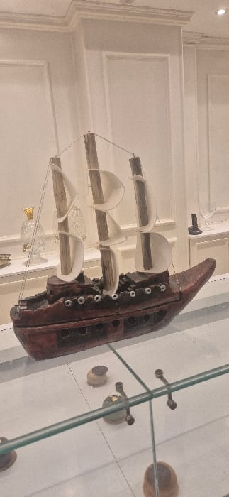 An antique shoe mold is on display in a glass case. The mold is crafted from dark wood and features metal eyelets. Alongside the mold, several white, curved forms are suspended by thin wires. These forms mimic the shape of a shoe's upper. The background features a white wall with classic paneling and a recessed ceiling with a light fixture. The glass case has decorative brass fixtures.
