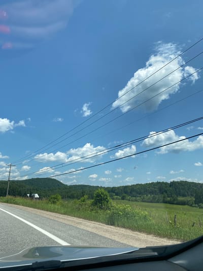 The image depicts a rural roadside scene under a bright blue sky with scattered white clouds. A paved road with a white line runs diagonally from the lower left to the middle right of the frame. To the right of the road is a grassy field with scattered wildflowers and a few wooden fence posts. Beyond the field, a treeline of green deciduous trees forms a gentle slope towards a distant, larger, forested hill. In the middle distance, nestled amongst the trees on the left side of the road, is a white building with a darker roof, possibly a barn or small house. Overhead, multiple power lines stretch across the sky, supported by a utility pole visible on the left side of the road. The perspective suggests the photo was taken from inside a vehicle, with the dashboard visible in the foreground. The environment is natural and undeveloped, characteristic of a countryside setting. There are no people or animals visible in the frame. The time of day appears to be daytime, with ample sunlight.