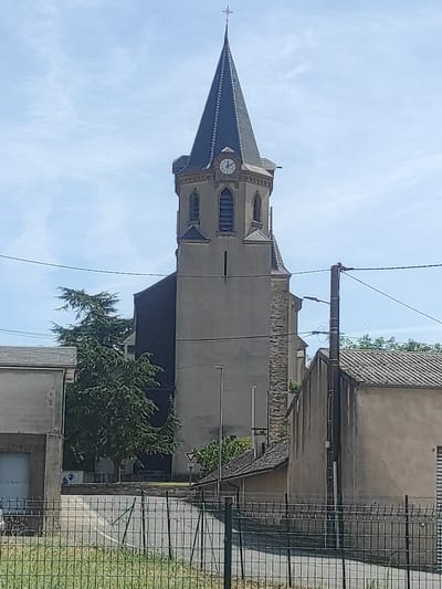 This is a vertically oriented, medium-shot photograph taken outdoors on a sunny day with a blue sky and scattered clouds. The main subject is a stone church tower with a dark, pointed roof and a white clock face. The tower has arched windows and decorative stonework. To the right of the church, a multi-story building with a tiled roof is visible, along with a chain-link fence and some greenery. The scene suggests a rural or small-town European setting, likely in Valence-d'Albigeois, France, given the context. There are no people visible in the image. The overall mood is peaceful and serene, with the bright sunlight casting shadows and highlighting the textures of the buildings.