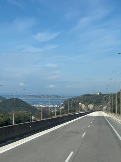 The image displays an elevated perspective of a multi-lane highway descending towards the coastal town of Αγία Μαρίνα, Greece, under a clear blue sky with minimal scattered cirrus clouds.
The foreground is dominated by a descending segment of an asphalt highway, presenting two marked lanes delineated by white dashed lines and solid white shoulder lines. A dark grey concrete barrier borders the left side of the road. The road surface is clear of vehicles or pedestrians.
Parallel rows of uniform, tall grey streetlights with horizontal luminaires line both sides of the highway, extending into the distance. The immediate environment comprises green, densely vegetated hillsides. Beyond the highway's left flank, a wide expanse of deep blue sea or bay is visible, with the small coastal settlement of Αγία Μαρίνα nestled along its shore, characterized by numerous light-colored structures. Distant islands or peninsulas are visible across the water, and a large white vessel is present within the bay.