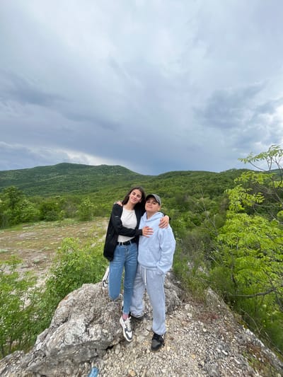 Two people are standing on a rocky outcrop overlooking a hillside covered in trees and shrubs. The foreground includes the gray rocks and vegetation, and in the distance, one can see the sky with cloudy skies. The location is likely near Kamchia, Bulgaria, given the image's description. The people are close together and it seems they are on a cliff, posing for a picture.