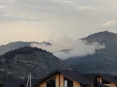 The image shows a cloudy sky over a mountainous landscape. In the foreground, the roofs of wooden houses are visible. The mountains are covered with trees and rocks. Some parts of the mountains are obscured by clouds. The overall impression is one of a natural, possibly remote, setting. The city of Anos, Russia is mentioned as the location.