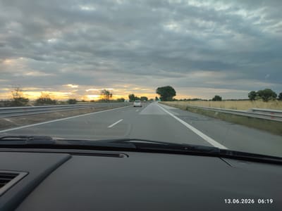 A first-person perspective shot shows a car driving on a highway at dusk. The sky is filled with dark, dramatic clouds with hints of sunset glow peeking through on the left side of the frame. The road ahead is wet, indicating recent rain, and is divided by white lane markings. A few cars are visible in the distance. Guardrails line both sides of the highway, separating it from grassy fields. The dashboard of the car is visible in the foreground. A timestamp in the lower right corner reads "13.06.2026 06:19". The image appears to be taken in Tsaratsovo, Bulgaria.