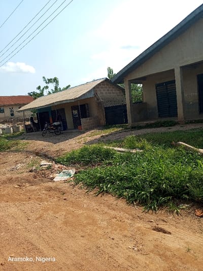 The image shows two structures in the city of Ilobu, Nigeria. The structures are simple, with a roof and walls made of concrete blocks. The larger structure has a porch. The smaller structure has a window and a door, and a motorcycle is parked in front of it. The two structures are separated by a patch of green grass, and a dirt road runs between them and the camera. In the foreground of the image is more green grass and dirt. The sky is blue with a few white clouds. The location of the image is in the city of Ilobu, Nigeria, based on the coordinates provided.