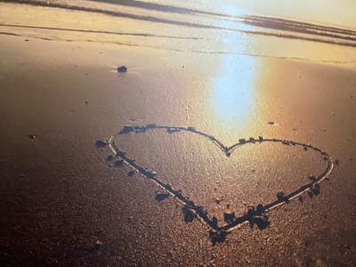 The image displays a close-up, low-angle view of a heart shape drawn into wet sand on a beach. The sand is dark and reflective, indicating recent water presence or saturation. Small, dark scattered pebbles and debris are visible on the sand surface. In the background, shimmering reflections of what appears to be a low sun or bright light source are evident on a body of water, possibly the ocean or a large lake, with horizontal wave lines visible further out. The overall lighting casts a warm, golden-orange hue across the scene, characteristic of sunrise or sunset. No individuals are present in the frame, nor are any objects other than the natural beach elements and the drawn heart. Given the depiction of a beach environment, this scene is not located in Lurgan, United Kingdom, as Lurgan is an inland town without a coastline.