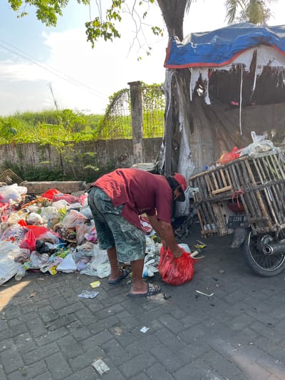 In Gedang, Indonesia, a man in a red shirt and camouflage shorts is picking through a large pile of garbage on a paved street. He holds a bright red plastic bag in his hands, appearing to be either adding to or sorting through the waste. The garbage includes numerous plastic bags, some filled and others empty, as well as what appear to be food wrappers and organic waste. To the right of the garbage pile is a motorcycle with a wooden cage attached to the back, covered with a blue and white tarp. The cage is also filled with various items, including plastic bags and what looks like food. A license plate with the number "B 6091 NY" is visible on the motorcycle. In the background, a concrete wall with overgrown vegetation can be seen, along with power lines and a bright, clear sky. The scene suggests a collection or sorting point for refuse.