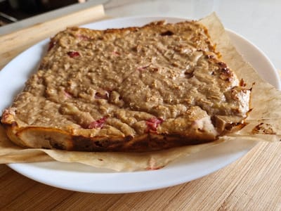 A rectangular baked item, light brown with a textured, uneven surface and visible reddish-pink fruit inclusions, rests on a crumpled piece of light brown parchment paper. This assembly is centered on a white, round ceramic plate. The plate is positioned on a light-colored wooden surface exhibiting horizontal grain patterns. The scene depicts an indoor environment located in the city of Niedernhausen, Germany. No individuals, actions, or interactions are present.