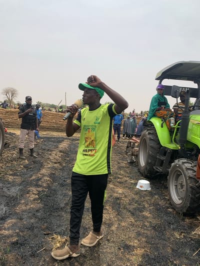 The image shows a man speaking into a microphone, holding a microphone in his right hand and with his left hand raised above his head. He is wearing a green shirt with a picture of a lion, black pants, and a green cap. He is standing in a field that appears to have been recently burned. In the background, there are other people, including a man in a security uniform standing with his arms crossed and a woman in a green hijab sitting in a green tractor. The tractor is parked in the background and the word "FAP" is visible on the side. This scene appears to be in a rural area in Ringimawa, Nigeria. The area is located in the vicinity of a farm or agricultural land. The man may be addressing the crowd or giving a speech. There are also other people standing around, likely listening to the man speak. The scene seems to be a gathering in a rural area. The coordinates provided are 11.666 latitude and 8.416 longitude.
