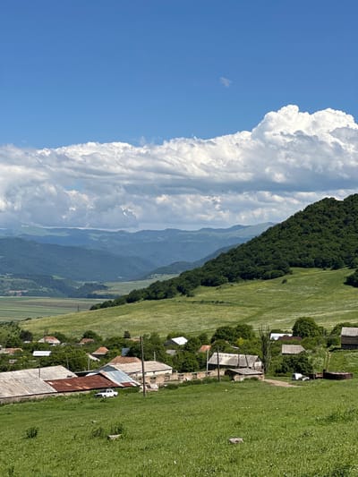 The image depicts a rural landscape with a village in the foreground, set against a backdrop of rolling green hills and distant mountains. The sky is a vibrant blue, dotted with large, fluffy white clouds. In the village, several houses with red and grey roofs are visible, surrounded by trees. A white car is parked in front of one of the houses. The ground is covered in lush green grass, with some scattered stones. The overall scene suggests a peaceful, rural setting in Aygehat, Armenia.