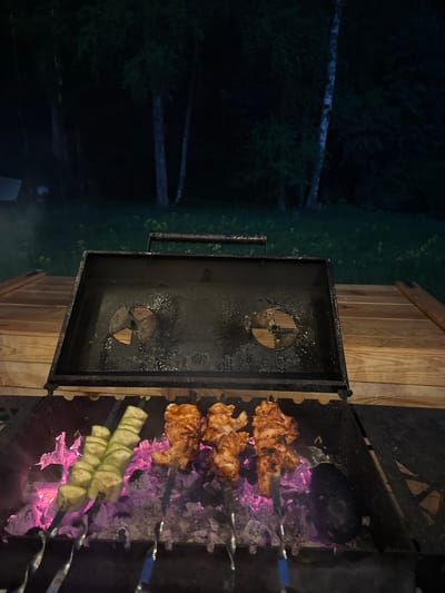 Image depicts a barbeque at night in Малеевка, Russia. Skewers with meat and zucchini are placed over hot coals in a black metal grill. A partially opened container attached to the grill is visible to the left. Wood and trees are visible in the background.