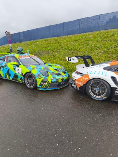 This image depicts two stationary Porsche 911 GT3 Cup race cars on wet asphalt, likely at a circuit within the city of Flatschach, Austria, consistent with the Red Bull Ring area. The environment is overcast with a grey sky.

On the left, a Porsche 911 GT3 Cup car, designated with number "5" and associated with driver "S. Spreng," features a camouflage-style livery in fluorescent green, black, and light blue. Visible sponsors include "DEKRA," "MOBIL1," "MICHELIN," and "TAG HEUER." A green and black racing helmet rests on the car's roof, alongside a clear plastic water bottle. A small, convex traffic mirror is mounted on a pole immediately behind this car, which also displays a white "WC" sign pointing left and a red circular "no stopping" sign.

The second race car, partially visible on the right, is also a Porsche 911 GT3 Cup. It displays a white base livery with orange and light blue accents. The word "ELLITE" is prominent in light blue on its side. Other visible sponsors include "bwh" and "Berger." This car features a large black rear wing with "Fellmayer Hachte" visible on the endplate. Both cars are equipped with Michelin tires.

The background consists of a sloping green grassy bank topped with a dark blue safety fence. No individuals are present in the frame.