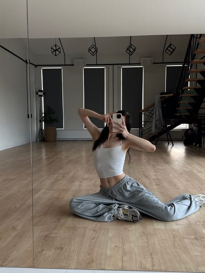 A person is taking a mirror selfie in a room with hardwood floors. The person is wearing a white tank top and grey sweatpants with athletic shoes. They are sitting with their legs extended. In the background, a staircase with a metal railing is visible. There are also windows with blinds, and some abstract geometric light fixtures hanging from the ceiling. A small plant in a basket is also in the background. The room appears to be a modern living space or studio. The lighting suggests it is daytime.