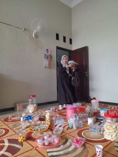 The image depicts an indoor scene within a building in Ketegan, Indonesia. The floor is covered by a patterned rug featuring interwoven brown, orange, and off-white geometric designs. Spread across the rug are numerous plastic containers holding food and beverages, including clear plastic drinking cups, various plastic pitchers, multiple clear jars filled with snacks such as white fried crackers and small yellow spherical items, and plastic trays containing individual dessert portions and cutlery. In the background, a woman stands positioned within a partially open dark brown wooden doorway. She wears a light-colored hijab and a long black dress, and is actively manipulating a white cloth object with both hands. A wall-mounted oscillating fan with visible wiring is fixed to the light gray wall on the left. Above the doorway, three dark rectangular vents are visible. A cardboard box labeled "AQUA" and a large clear jar of snacks are visible on the floor in the left background. A small purple wall-mounted organizer with hanging items is visible to the left of the doorway.