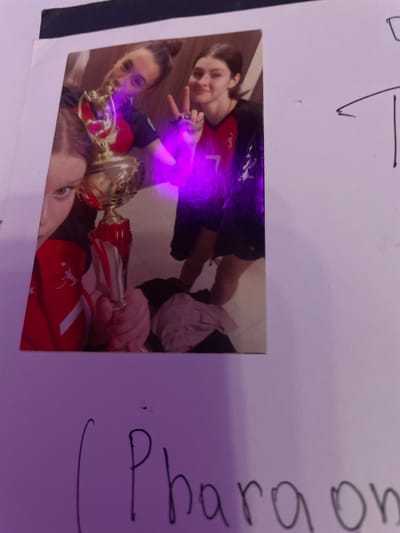 In the city of Vaganovo Massiv, Russia, a photograph depicts three individuals, likely young women, posing with a trophy. One individual, on the left, holds the trophy, their face partially obscured. Another individual in the center looks towards the camera with a hand raised in a peace sign. The third individual, on the right, also smiles at the camera, their arm around the central person. They appear to be wearing athletic attire, specifically jerseys with red and black accents. The trophy is gold with red accents. The background is indistinct, but the image is illuminated with a purple light. Below the photograph, handwritten text is visible.