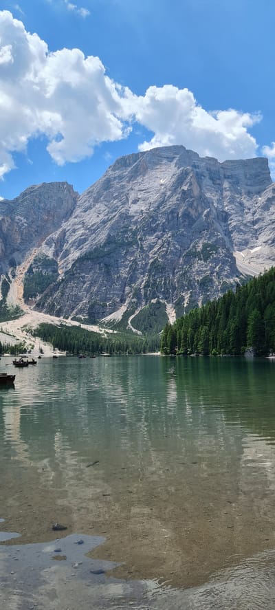 A vertical, eye-level shot captures the serene beauty of Lake Braies, also known as Pragser Wildsee, in Prags - Braies, Italy. The lake's clear, shallow waters in the foreground reveal a sandy bottom dotted with rocks and submerged objects. Ripples gently disturb the surface, reflecting the sky and surrounding landscape.

In the middle ground, several wooden rowboats are scattered across the lake. Some are empty, while others are occupied by people who appear to be rowing or enjoying the scenery. The lake transitions to deeper, greener waters further from the shore.

A dense forest of tall evergreen trees lines the right bank of the lake, creating a vibrant green contrast against the rocky mountains in the background. The mountains dominate the upper portion of the image, their rugged, grey faces etched with ravines and patches of sparse vegetation. A few remnants of snow are visible on the higher slopes.

The sky above is a bright blue, adorned with scattered, fluffy white clouds that cast soft shadows. The overall scene is one of natural tranquility and majestic alpine scenery.