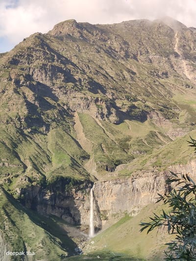 The image shows a panoramic view of a mountainous landscape with a waterfall cascading down a steep cliff. The mountains are rugged and rocky, with patches of green vegetation growing on the slopes. The waterfall is a prominent feature, with a thin stream of white water falling from a considerable height. The sky is partly cloudy, with some sunlight illuminating the scene. In the lower-left corner of the image, the text "deepak tha" is visible, suggesting it might be a caption or watermark. This scene is located in Yamling, India.