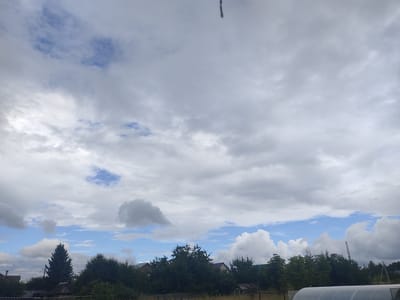 The image captures a cloudy sky over a rural area, possibly the village of Kamayka, Russia. The sky is a mix of white and gray clouds, with patches of blue sky visible. Below the sky, there is a dense line of trees, with some houses and structures peeking through. In the foreground, there are glimpses of a fence and a greenhouse-like structure. The overall impression is of a serene, overcast day in a countryside setting.