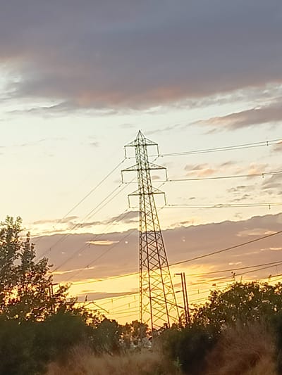 The image shows a pylon against a cloudy sky at sunset. The sky is illuminated with warm tones of orange and yellow, with darker clouds overhead. The pylon is made of metal lattice and is silhouetted against the bright sky. Power lines stretch horizontally across the frame, supported by the pylon. Trees and vegetation are visible in the foreground, with some of them illuminated by the setting sun. The overall scene depicts a rural or semi-rural landscape at dusk, with the presence of industrial infrastructure. The location is specified as Somaglia, Italy.