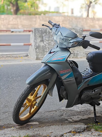 A silver-grey Yamaha Jupiter MX motorcycle is prominently parked on the side of a paved road under daylight conditions. The motorcycle, positioned at a slight angle facing left, is equipped with striking gold-colored front wheel rims. The main body features blue and red accents, with the "YAMAHA" logo clearly visible in red and blue text on the side panel. Below it, the text "DiASiL" and "JUPITER MX" (partially visible) are also present. A black seat and a small chrome luggage rack are visible on the rear section, while a black bag or cover hangs near the foot area. The motorcycle is resting on its kickstand.
The setting appears to be an outdoor urban or suburban street, consistent with Nam Nha Trang, Vietnam. In the mid-ground, a low concrete barrier with red and white striped markings runs horizontally, separating the immediate roadside from a deeper background. The background is softly blurred, revealing more paved road, concrete structures, and lush green foliage, suggesting trees and other greenery under bright, natural light. Scattered yellow petals are visible on the ground near the motorcycle's front wheel and alongside the curb. No people are visible in the image.