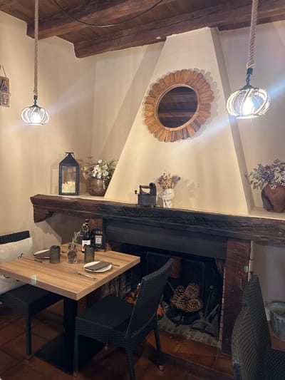 This image depicts the interior of a restaurant in Patones De Arriba, Spain, likely a cozy dining area near a fireplace.
The scene is dominated by a large, rustic wooden fireplace mantel. Above the mantel, a round wooden mirror with a sunburst design hangs on a cream-colored wall. Two decorative pendant lights with rope detailing hang from the wooden beam ceiling, one on the left and one on the right of the mirror.
On the mantelpiece, a black metal lantern with three lit candles is positioned on the left. To the right of the lantern, a vase containing a bouquet of white and purple dried flowers sits. Further to the right, a small, white ceramic bust with a flower crown holds another small arrangement of dried flowers, and a vintage black iron is visible to its left. On the far right of the mantelpiece, a large terracotta pot holds a bouquet of purple and white dried flowers.
In the foreground, a square wooden table is set for two. Each place setting includes a dark-colored plate, cutlery, and a dark grey tumbler. In the center of the table, a small vase holds a sprig of rosemary and a few small objects. Two wine bottles and a QR code menu stand behind the vase. Two black woven chairs are placed at the table, one on the left and one on the right. Another black woven chair is partially visible on the far right of the image.
The fireplace itself is dark, and a metal grate containing several large pinecones is visible within it. The floor in front of the fireplace appears to be tiled with terracotta-colored tiles. A bucket with some material in it is visible on the far right. A white cushion with a black pattern is placed on a seating area to the left of the table.