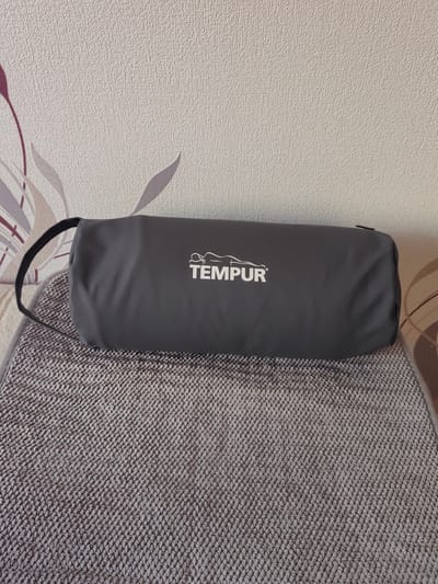 A gray cylindrical bag with a white logo is placed on a textured gray mat. The bag has a black strap attached to its left side. The logo on the bag features a stylized human figure lying down, with the word "TEMPUR" underneath. The background consists of wallpaper with a subtle textured pattern and floral motifs in shades of beige, purple, and gray. The scene is indoors, likely in a residential setting. No people are visible in the image.