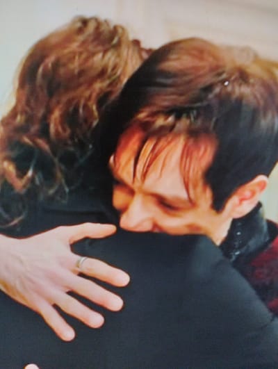 This is a close-up, over-the-shoulder shot of two individuals embracing. The person in the foreground, facing away from the camera, has short, dark hair with some reddish highlights and is wearing a dark-colored jacket with red accents. Their left arm is visible, resting on the back of the other person, and a ring is present on their left ring finger. The person in the background, whose face is partially obscured by the foreground individual, has long, curly, light brown hair and is also embracing the other person. The background is a plain, light-colored wall, suggesting an indoor setting. The image quality is pixelated, indicating it was likely taken from a screen display. There is no indication of the city of Yangibazar, Uzbekistan within the visual content of the image.