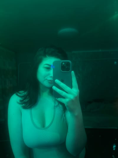 A person is taking a selfie in a dimly lit room with green lighting. The person is holding a smartphone to take the photo, and their face is partially obscured by the phone. They are wearing a light-colored sports bra. The room appears to have a mirror and some furniture in the background, though it is difficult to discern details due to the lighting. The image does not contain any information about the location in the city of Gavrilov Posad, Russia.