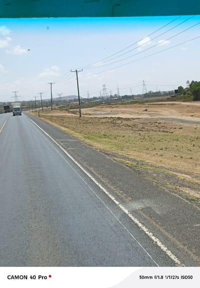 This image depicts a wide-angle view of a highway in Hells Gate ward, Kenya, captured from within a vehicle. The asphalt road occupies the left foreground and mid-ground, featuring a solid white line delineating the right edge of the main visible lane. Adjacent to the road is a gravel shoulder, transitioning into an expanse of dry, sparse grassland and exposed soil extending to the horizon.
In the mid-ground, utility poles with power lines run parallel to the road. Further into the background, numerous large high-voltage electricity pylons are visible against a backdrop of distant hills and an expansive, clear blue sky with minimal cloud cover. Scattered structures and light industrial elements are discernable in the far distance beneath the pylons.
Two vehicles are visible on the road in the distance, moving away from the viewer: a white box truck or similar medium-sized truck, followed by a darker, larger truck. No pedestrians or other discernible human figures are present. The top portion of the image shows a blue-tinted obstruction, consistent with a vehicle's windshield visor or tint. Camera metadata, "CAMON 40 Pro" and "50mm f/1.8 1/1727s ISO50," is present at the bottom of the frame.