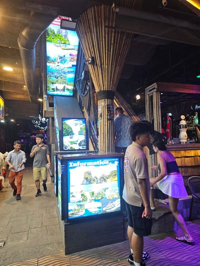 This media file depicts an outdoor scene in Ban Klong Haeng, Thailand, likely at night or in the late evening due to the artificial lighting. The setting appears to be a bustling entertainment area, possibly a bar or restaurant, with a prominent use of bamboo in its construction, seen in the overhead structures and wall paneling.

Several individuals are visible. In the foreground, two men are standing on a stone-paved walkway. One man wears a light blue button-down shirt and orange pants, while the other is in a grey t-shirt and khaki shorts, holding a paper. Behind them, a young person with dark hair, wearing a white skirt and a dark top, is interacting with another individual whose face is not clearly visible. Further back, a person in a blue shirt stands near a bamboo structure.

The scene is illuminated by a mix of colored lights, creating a vibrant and energetic atmosphere. A large, bright LED screen displays a collage of tropical island scenery, featuring turquoise waters, lush green cliffs, and boats. Text is visible on this screen, including the word "Information" in English and possibly Thai script. Another smaller screen to the left also displays similar tropical imagery. In the background, glimpses of other establishments with neon signs and bar equipment, like beer taps, suggest a lively nightlife. The overall impression is one of a tourist-oriented destination, possibly a night market or a lively street.
