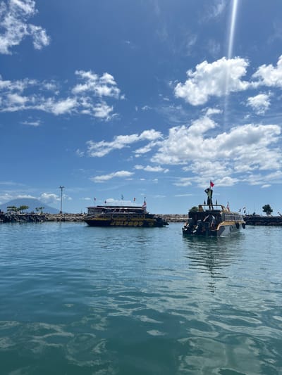 An eye-level medium shot captures a bright, sunny day at a harbor in Tidore Islands, Indonesia, with calm, blue water reflecting the sky and clouds. In the mid-ground, a long, dark boat with yellow lettering, possibly advertising "ZULFAD," is docked or moving slowly. To the right, a smaller, yellow-and-white boat with an orange trim is positioned closer to the viewer. This boat has several people on it, one standing at the stern holding a red flag. Another boat of similar design is visible behind it, further into the distance. The background features a rocky shoreline with scattered debris and a few trees, leading to a mountain in the far left. A lamppost stands near the shore. The sky is a vibrant blue, dotted with scattered white clouds, and a strong beam of sunlight cuts diagonally from the top right corner.