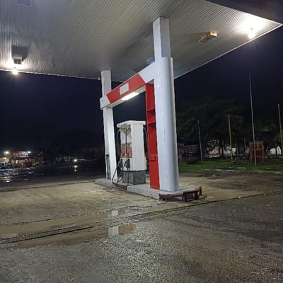 The image captures a quiet gas station at night in Kecamatan Kapongan, Indonesia. The primary subject is a fueling station structure with a large, flat gray canopy supported by prominent white and red pillars. Underneath the canopy, a white and red fuel dispenser stands, equipped with multiple nozzles and hoses, ready for use.

The ground around the dispenser and stretching into the foreground is a rough, unpaved or heavily worn asphalt/concrete surface. It appears wet, with numerous puddles reflecting the artificial lights from the canopy, suggesting recent rain. Visible tire tracks crisscross the damp ground. A small, dark wooden bench or step is placed near the base of one of the main support pillars.

In the background to the left, several small, modest structures, possibly roadside stalls or eateries, are dimly lit, with a dark silhouette of a mountain or large hill beyond them. To the right, trees, utility poles, and some indistinct brown structures are visible against the dark night sky.

No explicit activity is taking place; the station appears deserted. The time is clearly night, with the only illumination coming from the lights under the gas station canopy and a few distant lights. There is no clearly legible text visible on the dispenser or any other part of the scene. The overall atmosphere is still and somewhat desolate, characteristic of a late-night or deserted roadside stop in a rural or semi-rural area.