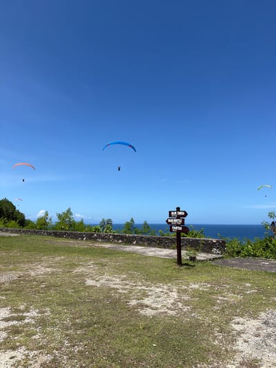 The image depicts a scene in Benoa, Indonesia, featuring paragliders in the sky. A brown wooden signpost stands near a grassy area, indicating directions to "Jalur Tangga," "Jalur Shuttle," and "Pantai." The sea and sky meet at the horizon, with a coastline visible. Several paragliders are scattered across the blue sky. A wall with greenery separates the grassy area from the coastal view.