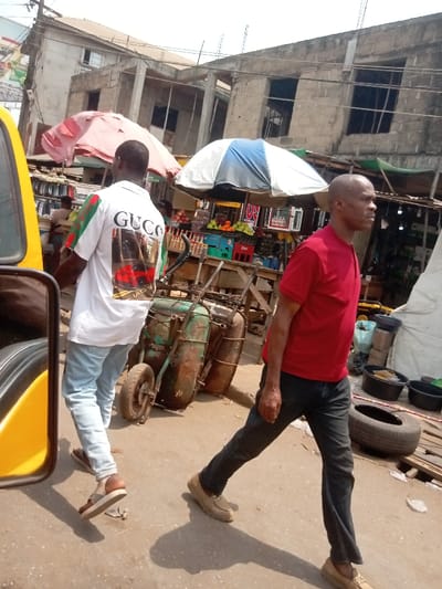 The scene depicts a sunny, outdoor street market environment in Umudioka, Nigeria. Two adult males are prominently featured. The first, on the left, has his back mostly to the camera, wearing a white short-sleeved polo shirt with "GUCCI" and a red and green stripe design, light blue jeans, and brown sandals. He is walking away from the viewer, glancing slightly to his right. The second male, on the right, is bald and walking in profile towards the right of the frame. He wears a red short-sleeved t-shirt, dark trousers, and light tan loafers, looking straight ahead.
Between the two individuals and slightly behind them, a rusty, green two-wheeled handcart or pushcart is visible, laden with cylindrical objects. In the background, market activity is evident under two open umbrellas: one red/pink and another white and blue striped. Under the blue and white umbrella, crates holding various fruits, including oranges and green produce, are displayed on a wooden stall. Further back, multi-story concrete buildings, some appearing unfinished with dark window openings, frame the street. Overhead, numerous electrical or utility wires crisscross the sky. The ground is light-colored dirt or pavement, showing prominent shadows from the individuals and objects, indicating direct sunlight. A partial yellow vehicle is visible on the far left foreground, likely a tricycle or bus. On the far right, a loose black tire and several black plastic basins are on the ground.